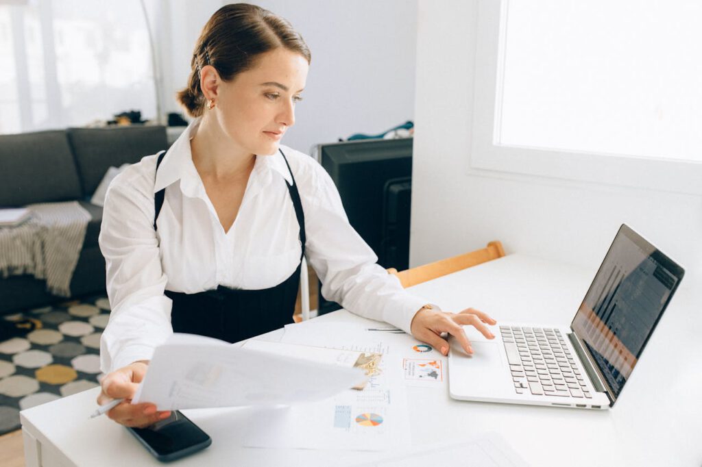 Woman at desk reviewing business reports on laptop, holding papers in a modern office setting.